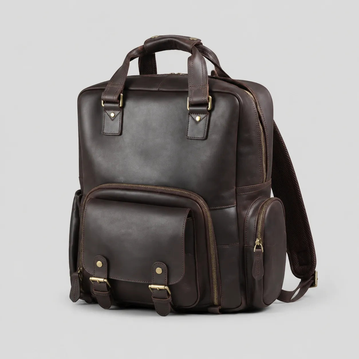 Dark brown Crazy Horse leather camera backpack on a light gray background, showing front view with top handle, front flap pocket with buckles, side pocket, and durable brass hardware. Suitable for DSLR cameras, laptop, and travel essentials.