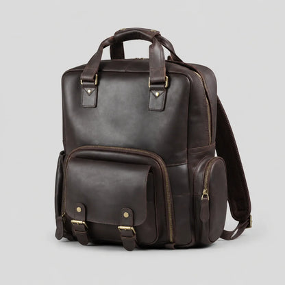 Dark brown Crazy Horse leather camera backpack on a light gray background, showing front view with top handle, front flap pocket with buckles, side pocket, and durable brass hardware. Suitable for DSLR cameras, laptop, and travel essentials.